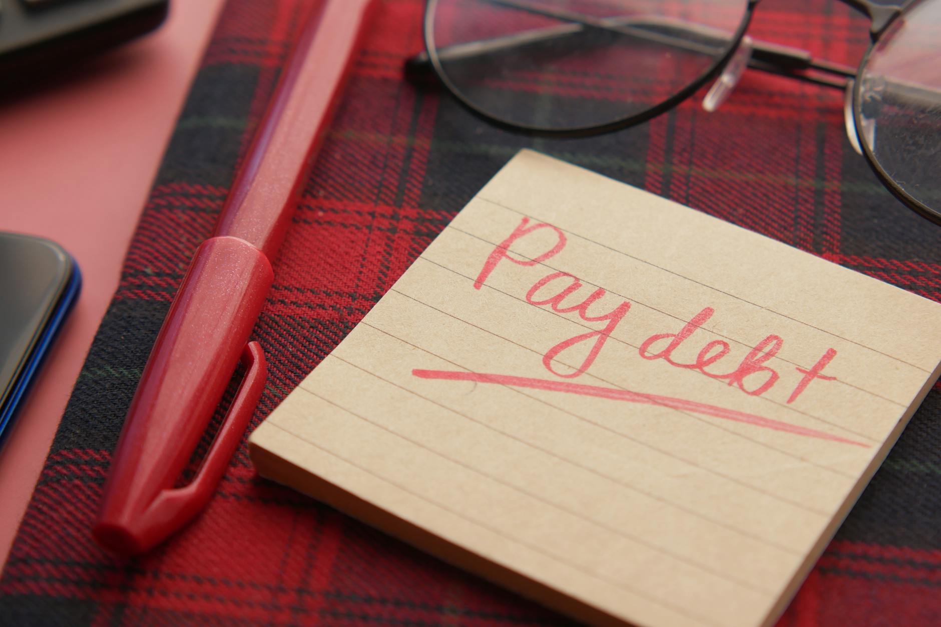 Kartka z napisem Pay debt i czerwony długopis na kraciastej tkaninie